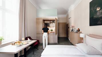 A hotel room with a bed, a desk with breakfast, a kitchenette, and a bathroom.