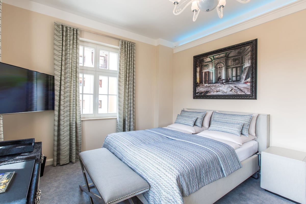 A bedroom with a bed, a flat-screen TV, a desk, and a large window with curtains.
