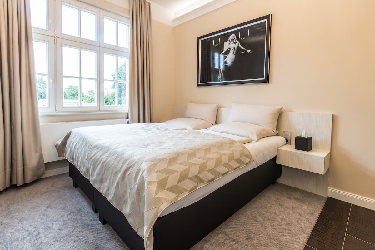 A bedroom with a bed, a nightstand, a framed picture, and a window with curtains.
