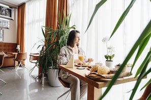 A person sitting at a wooden table with a laptop, a glass of orange juice, and a plate of food. There is a large potted plant and a small table with a white flower. The room has a modern design with a sofa and chairs.