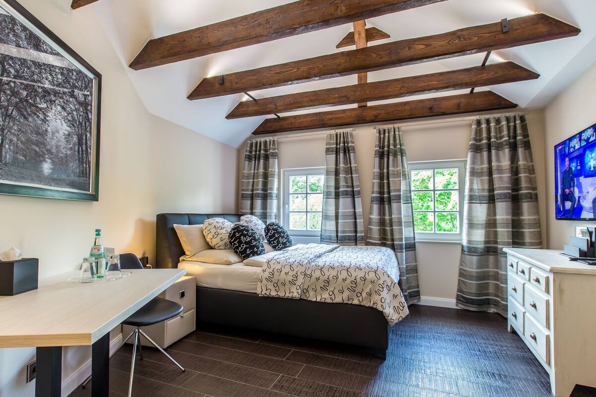 A bedroom with a bed, a desk, a chair, a TV, and a window with curtains.
