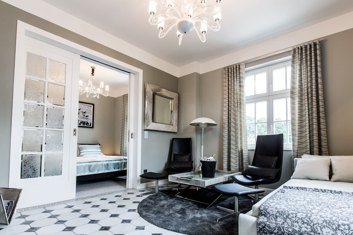 A hotel room with a bed, a desk with a chair, a lamp, a chair, a large window with curtains, and a chandelier.
