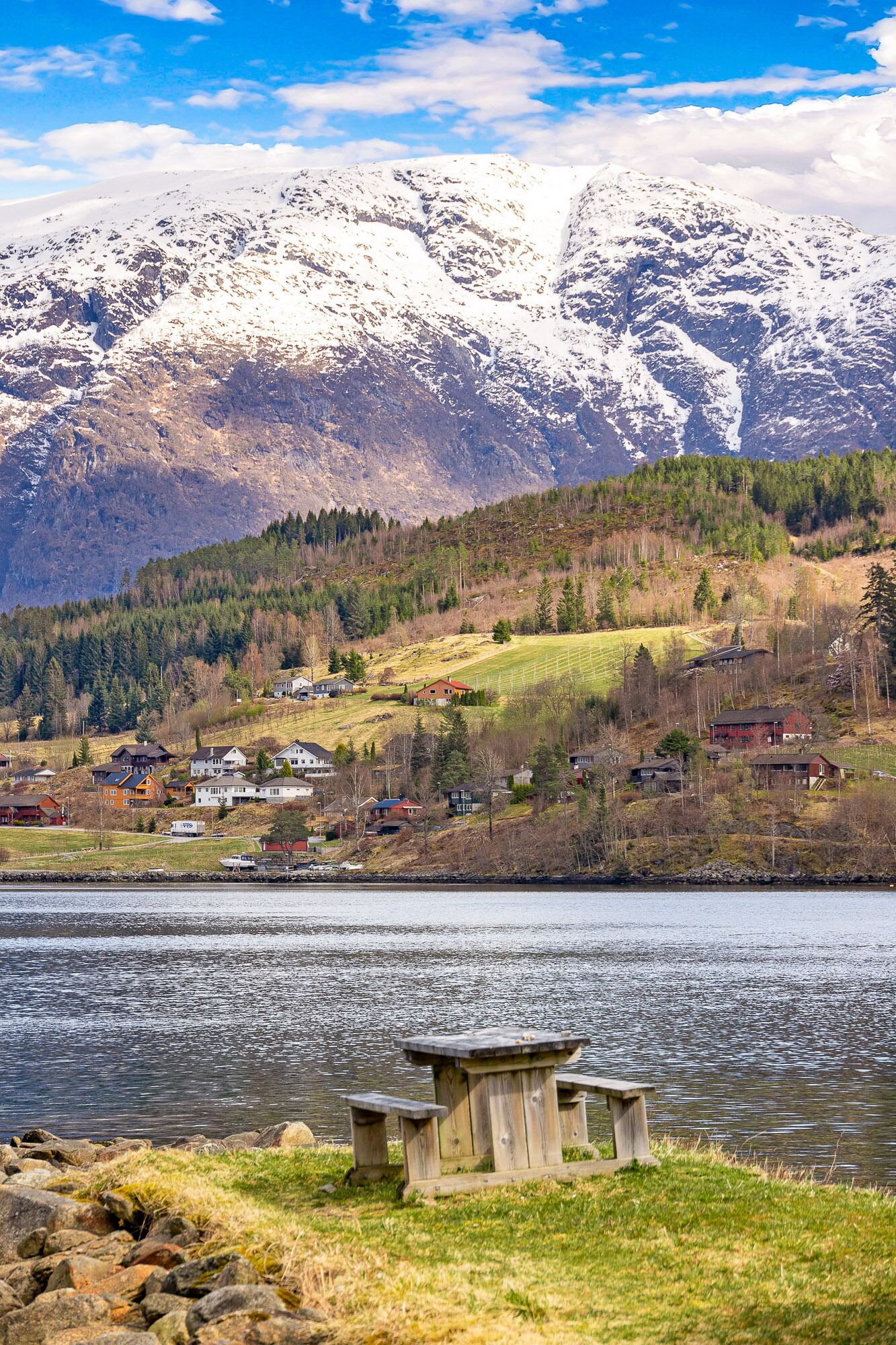 A lakeside view with a wooden bench, houses, and snow-capped mountains in the background.