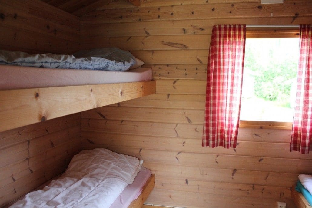 A small cabin with two bunk beds, a window with red and white checkered curtains, and a wooden interior.