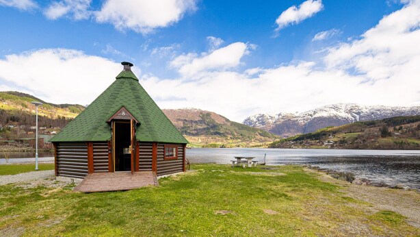 A small wooden cabin with a green roof, situated by a lake with mountains in the background. - Ulvik Camping (Ulvik)