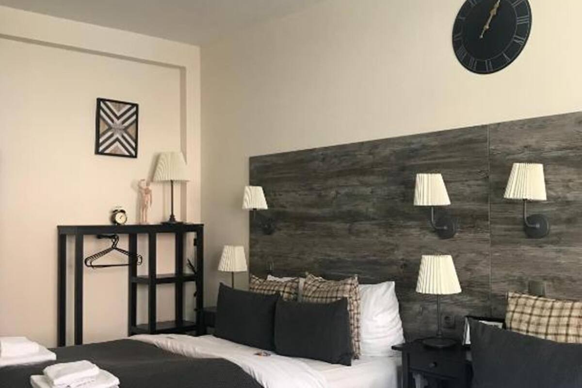 A hotel room with two beds, a wooden headboard, bedside lamps, a wall clock, and a small table with a lamp and a decorative item.
