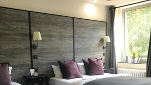 Habitación de hotel con dos camas, cabecero de madera, lámparas de noche y ventana con vistas a la vegetación.