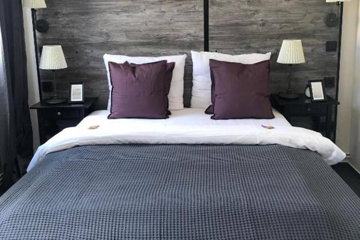 A neatly made bed with a grey textured bedspread, two white pillows, and two purple accent pillows. There are two bedside tables with lamps and a dark headboard.