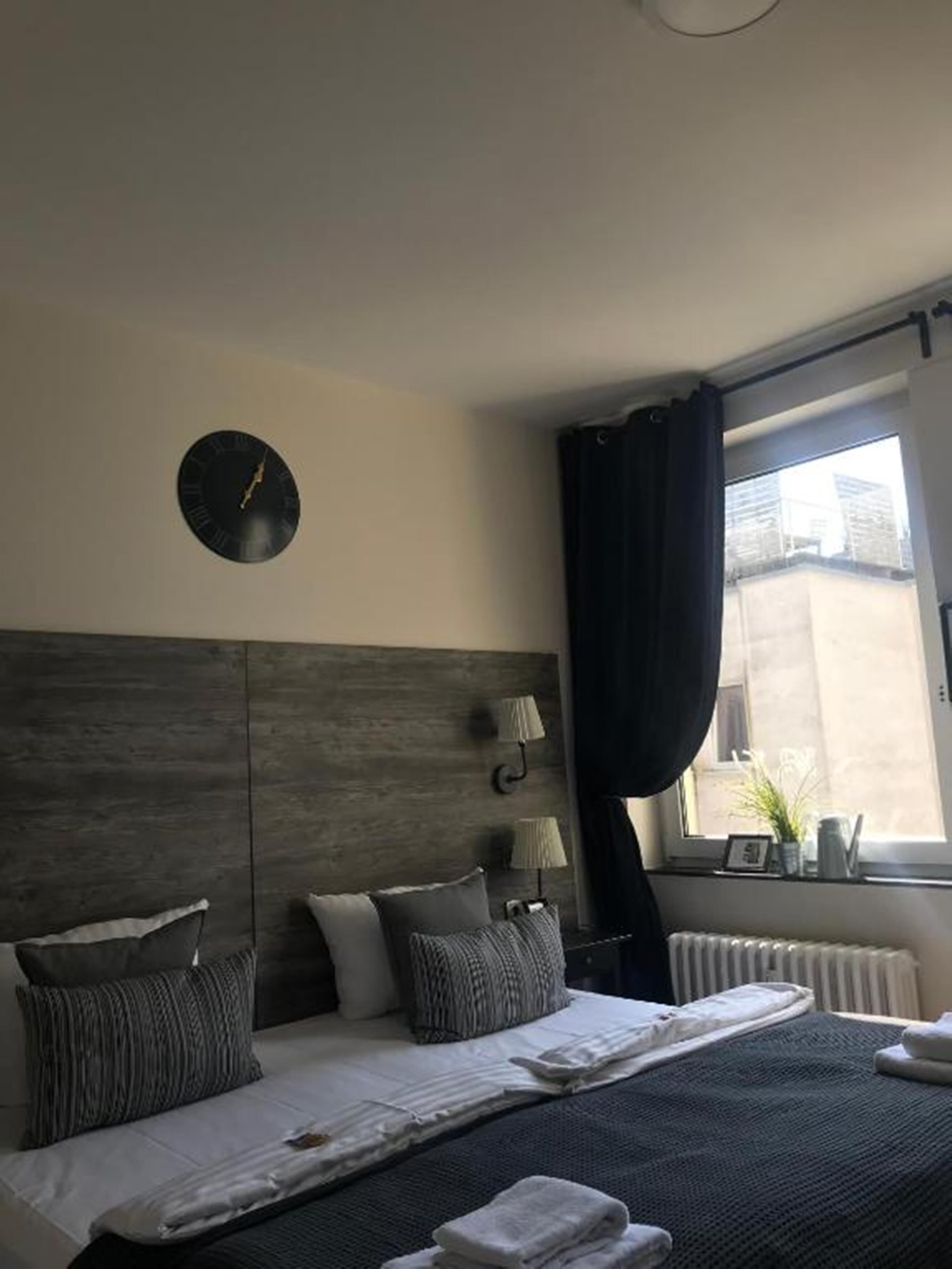 A modern bedroom with a wooden headboard, a round wall clock, two bedside lamps, a large window with curtains, and neatly folded towels on the bed.