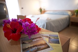 Een hotelkamer met een bed, een houten tafel met folders en een bloemenboeket.