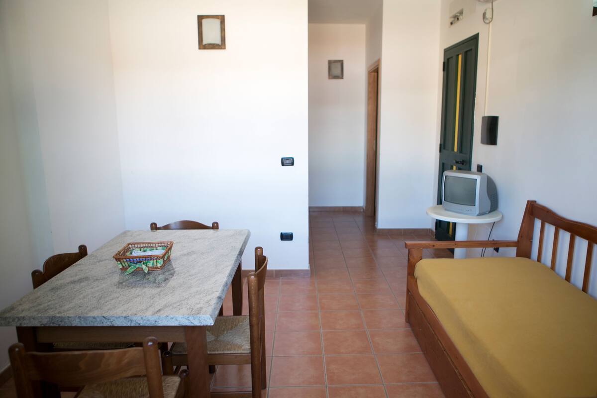 A dining area with a table and chairs, a small television on a side table, and a bed with a yellow mattress.