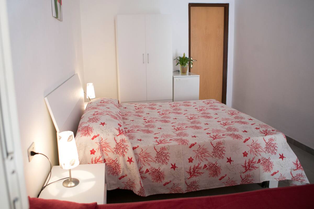 A bedroom with a bed, bedside table, lamp, wardrobe, and a small desk with a plant.