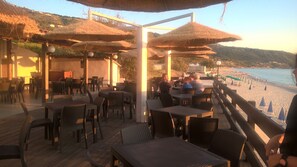 An outdoor dining area with tables and chairs, a wooden deck, and a view of the beach and ocean. - Villaggio Borgo Marino (Zambrone)
