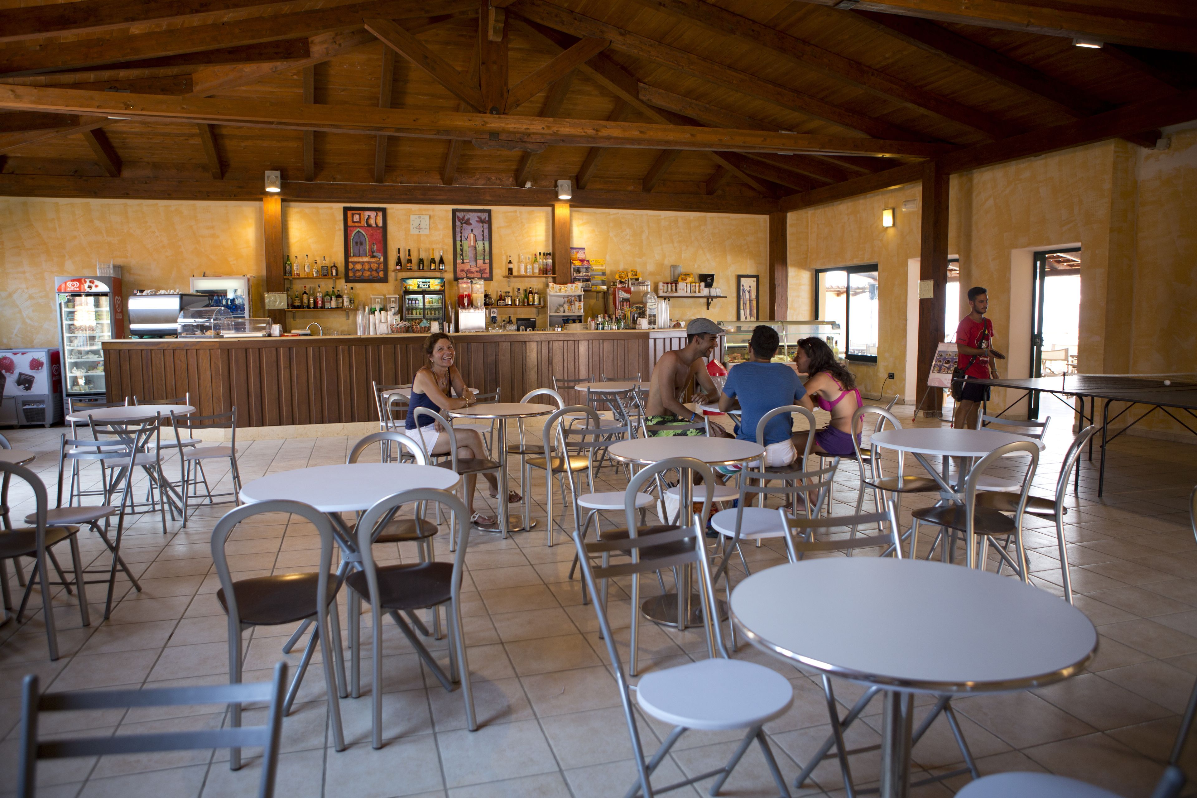 Un grand espace de restauration intérieur avec des tables et des chaises en métal, un comptoir de bar avec diverses bouteilles et un appareil à café.