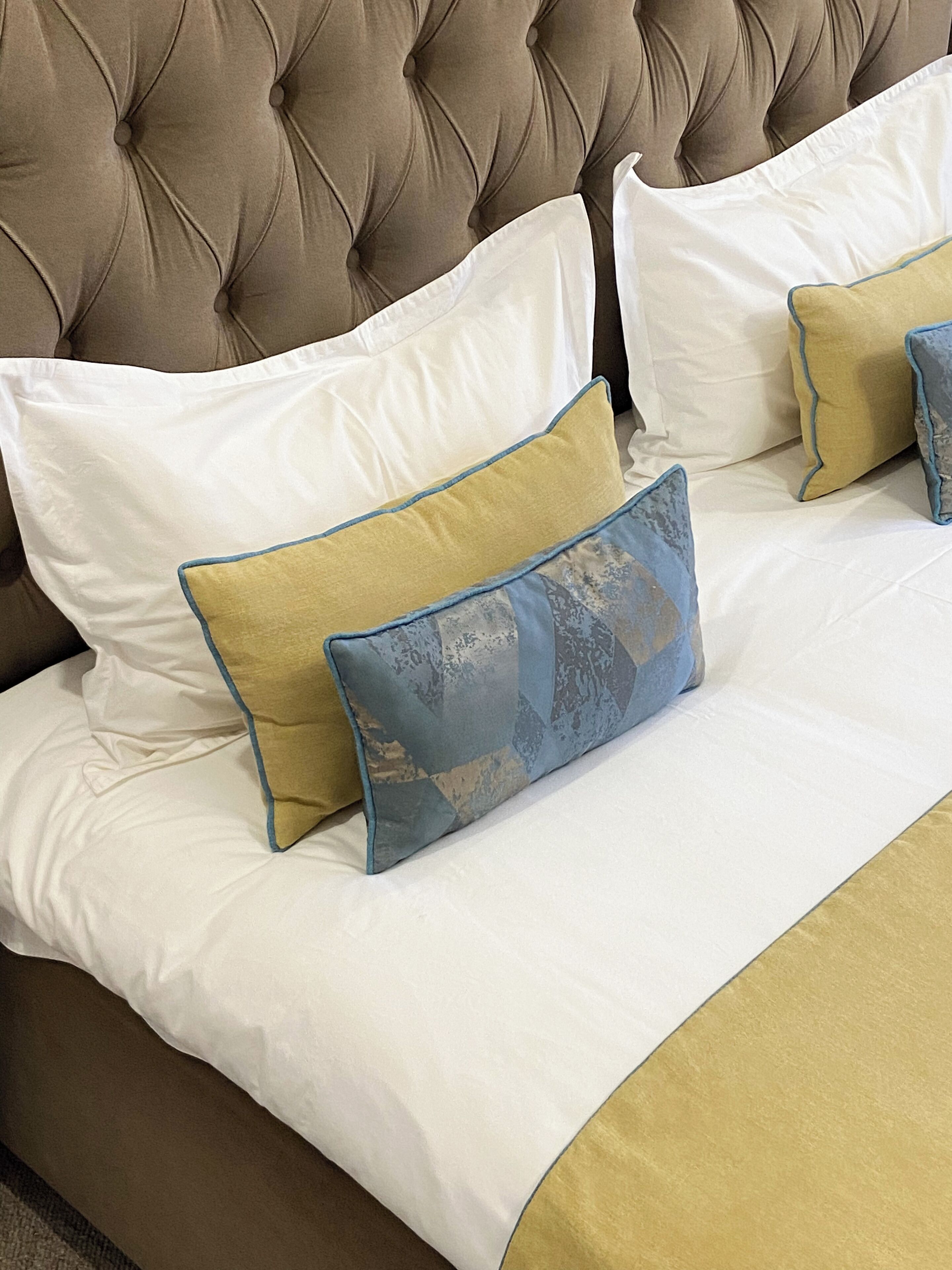 Una cama con cabecera capitonada, ropa de cama blanca y dorada, y almohadas decorativas.