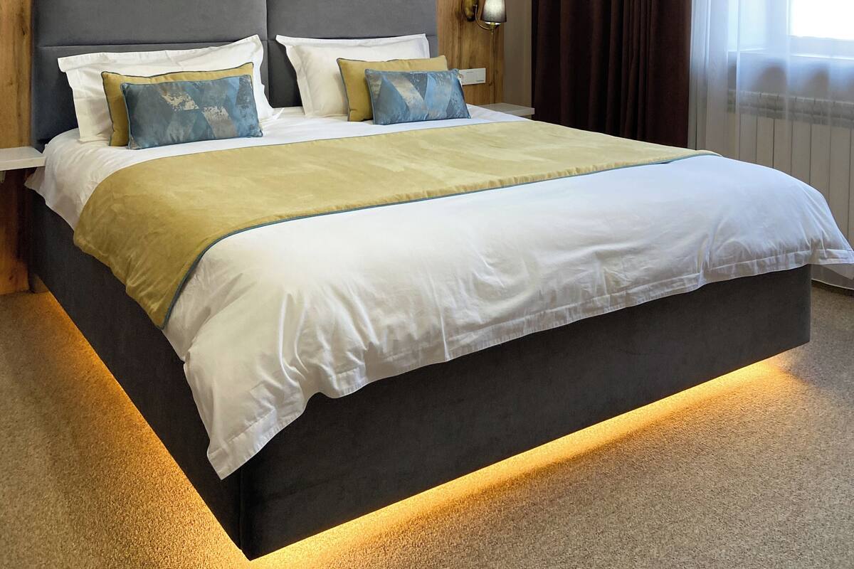 A hotel room with a large bed, a wall-mounted air conditioner, a bedside table with a lamp, and a window with curtains.