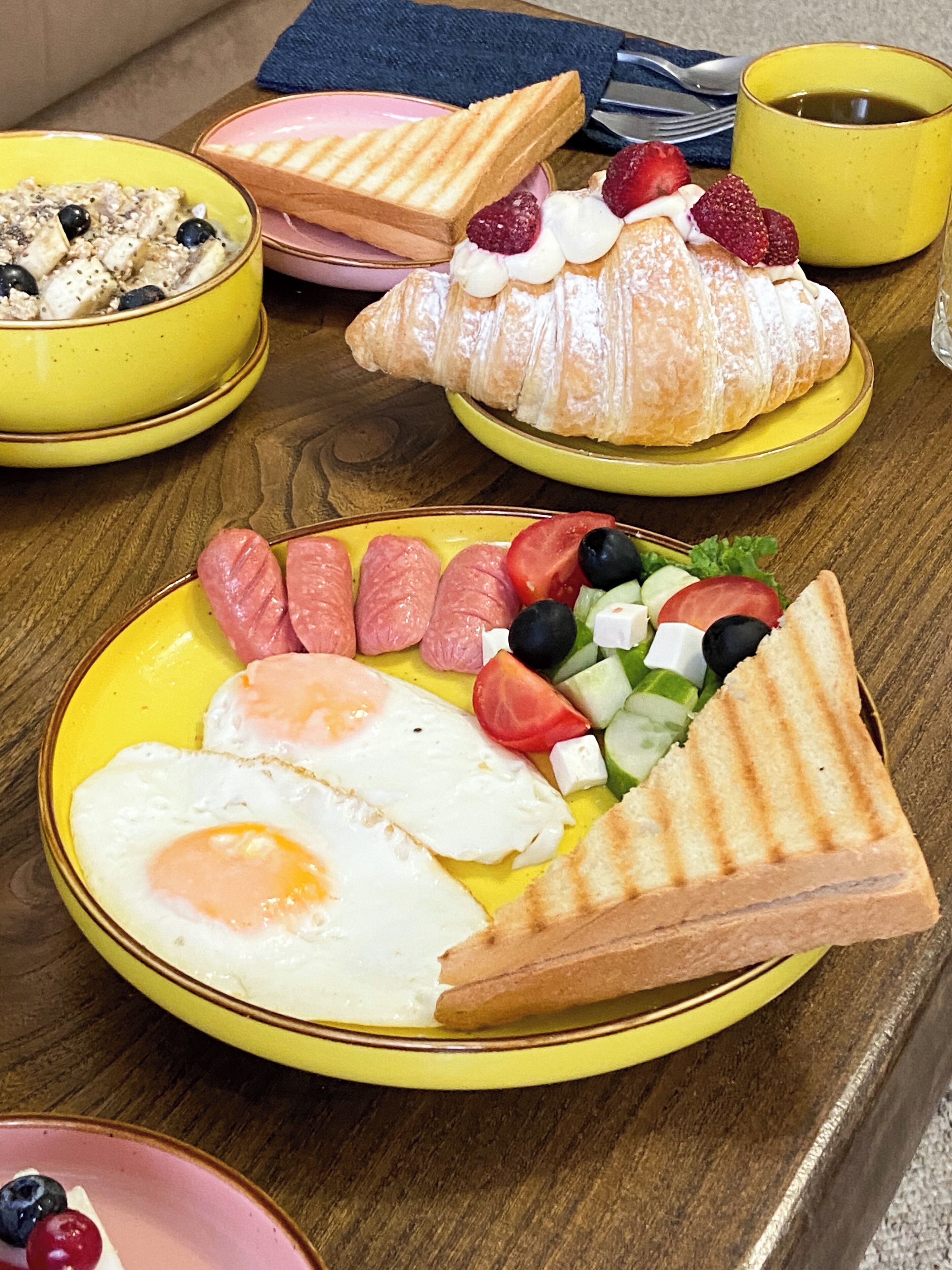 A breakfast table with plates of food including eggs, sausages, tomatoes, cucumbers, and a croissant.
