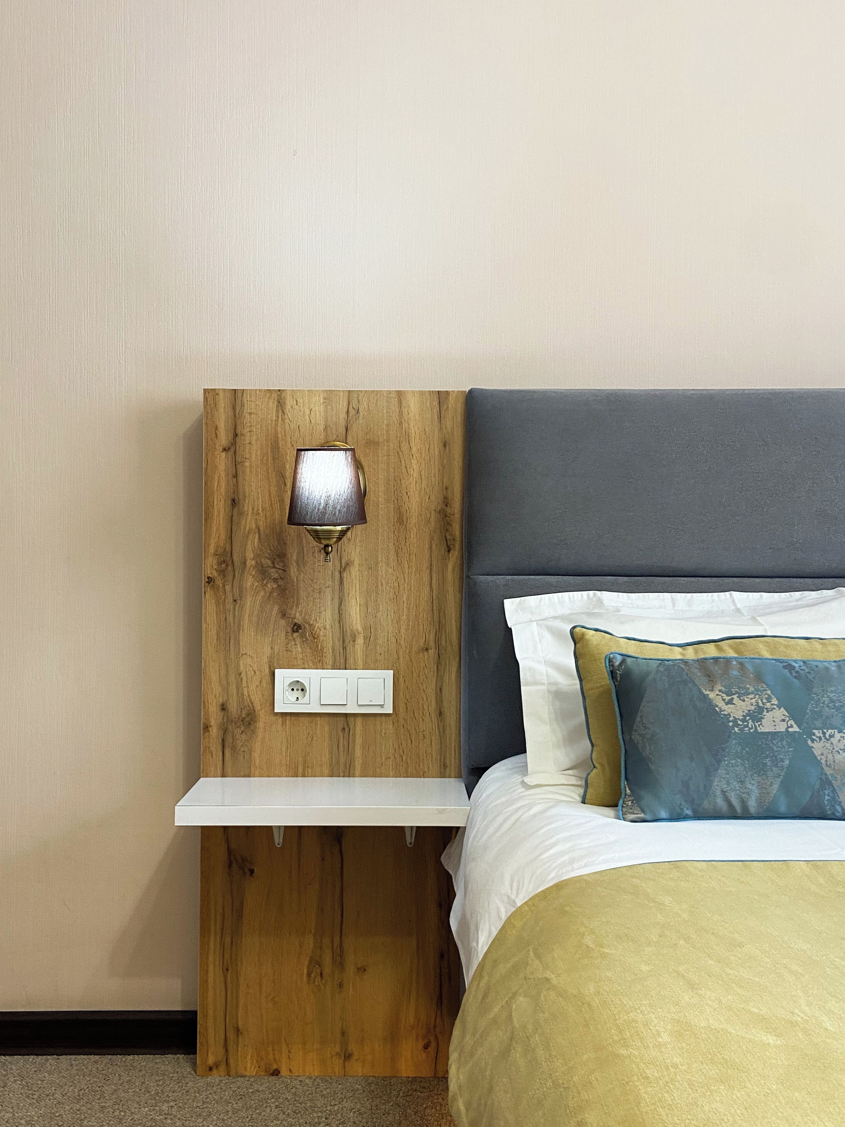 A bedroom with a wooden nightstand, a lamp, a bed with a grey headboard, and a white pillow.