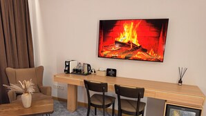 A hotel room with a long wooden desk, two chairs, a sofa, a coffee machine, a TV displaying a fireplace, and a minibar. - Keremet Hotel (Almaty)