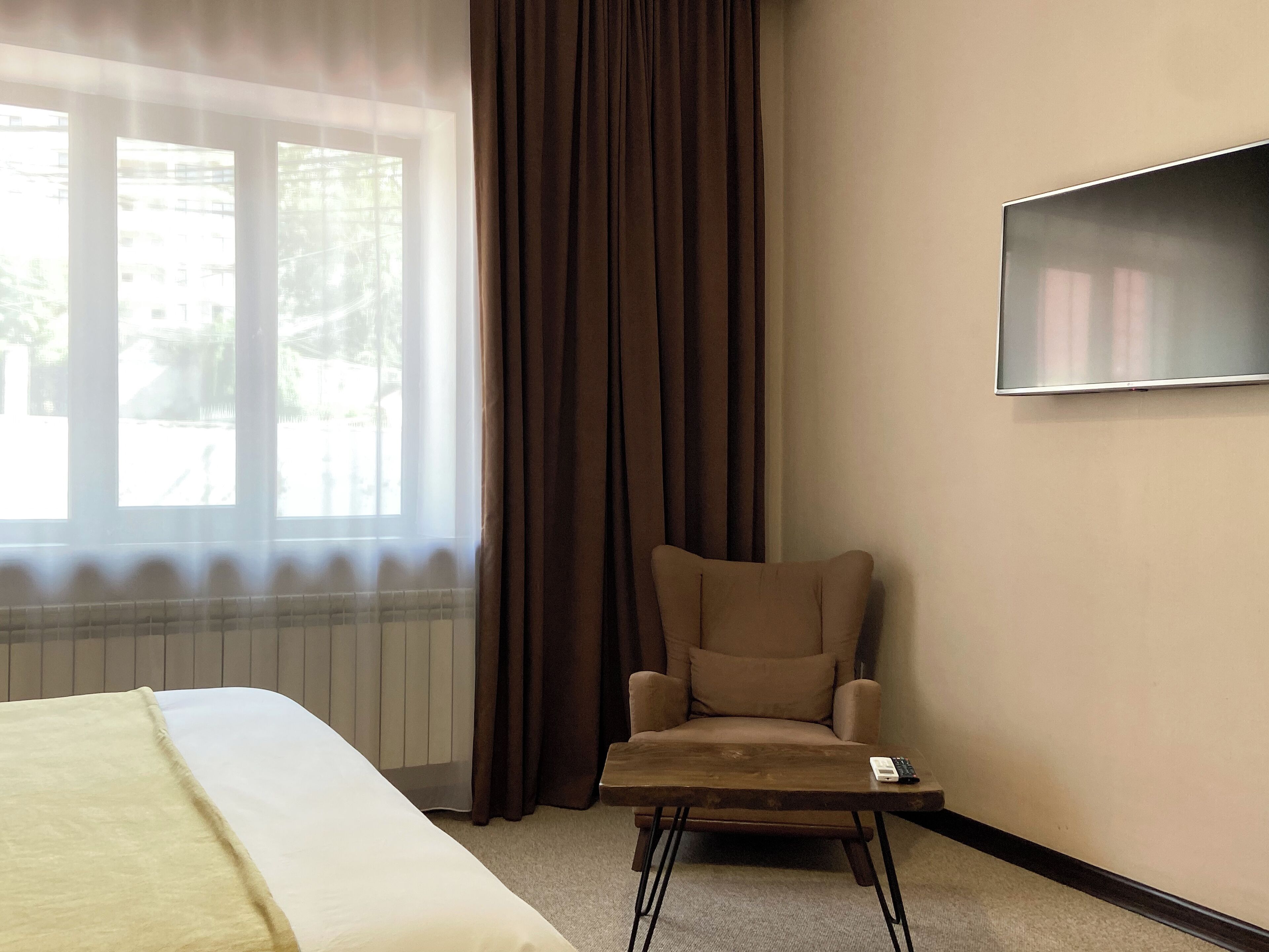 A hotel room with a bed, a chair, a small table, a TV, and a window with curtains.
