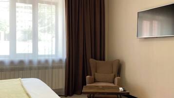 A hotel room with a bed, a chair, a small table, a TV, and a window with curtains.