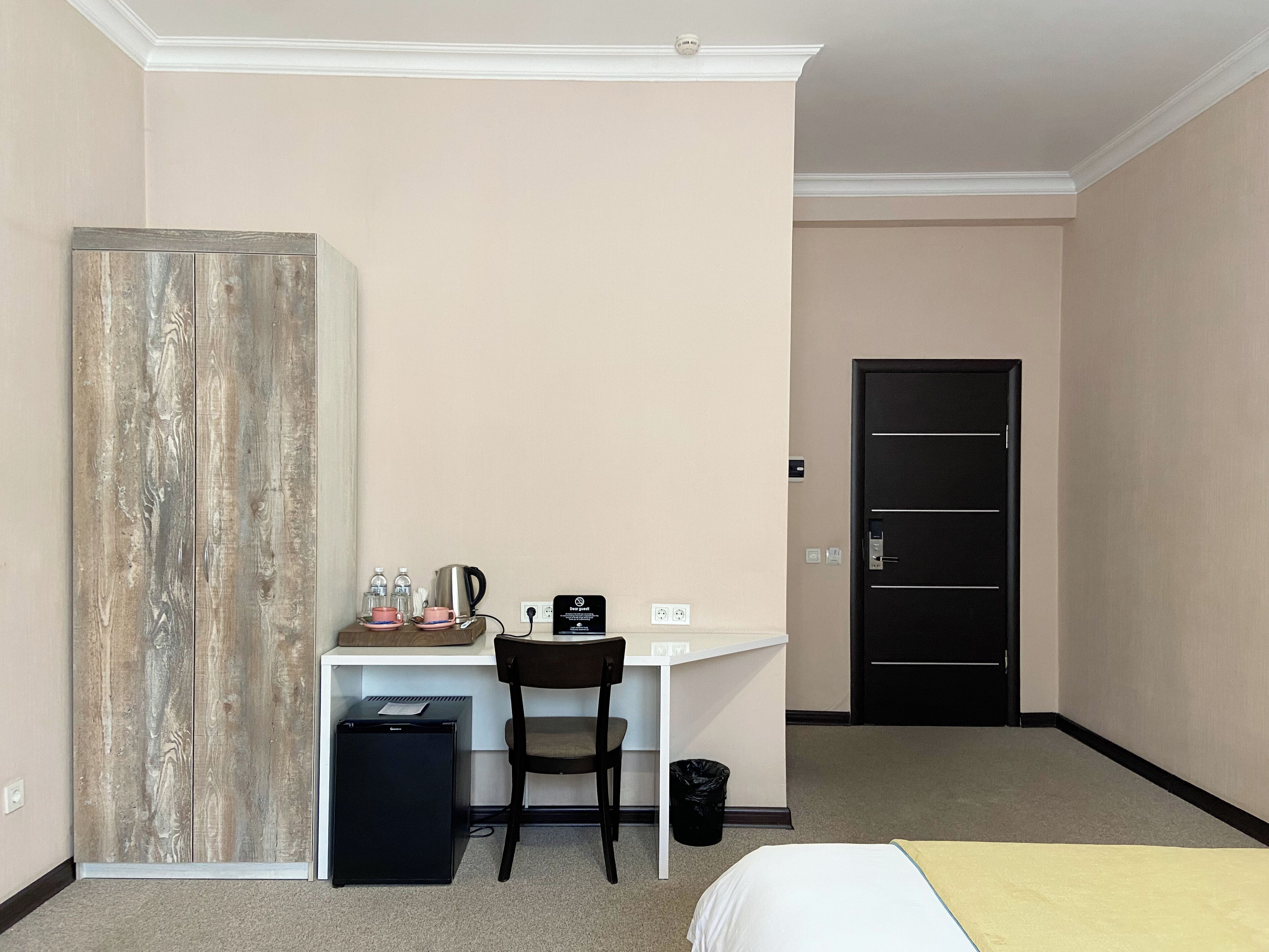 A hotel room with a bed, a desk with a chair, a small refrigerator, a trash bin, and a door.