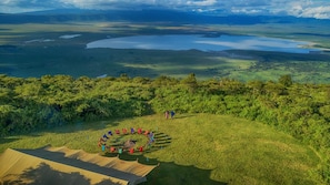 A campsite with tents and a circle of colorful flags, surrounded by lush greenery and a vast landscape with water bodies. - Pakulala Safari Camp - East Africa Camps (Ngorongoro)