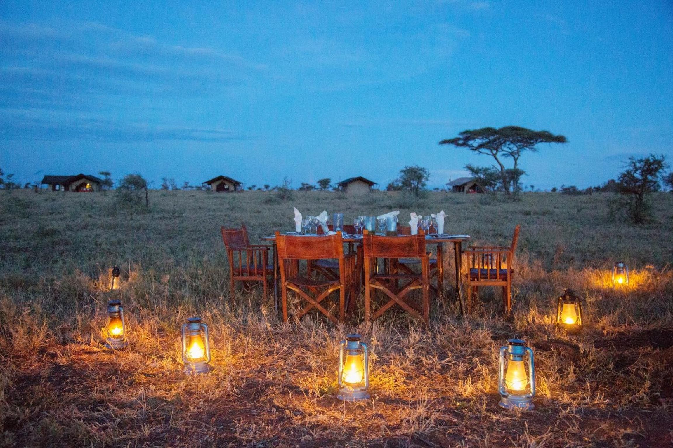 Mesa al aire libre con sillas y faroles iluminados, rodeada de hierba seca y un cielo despejado.