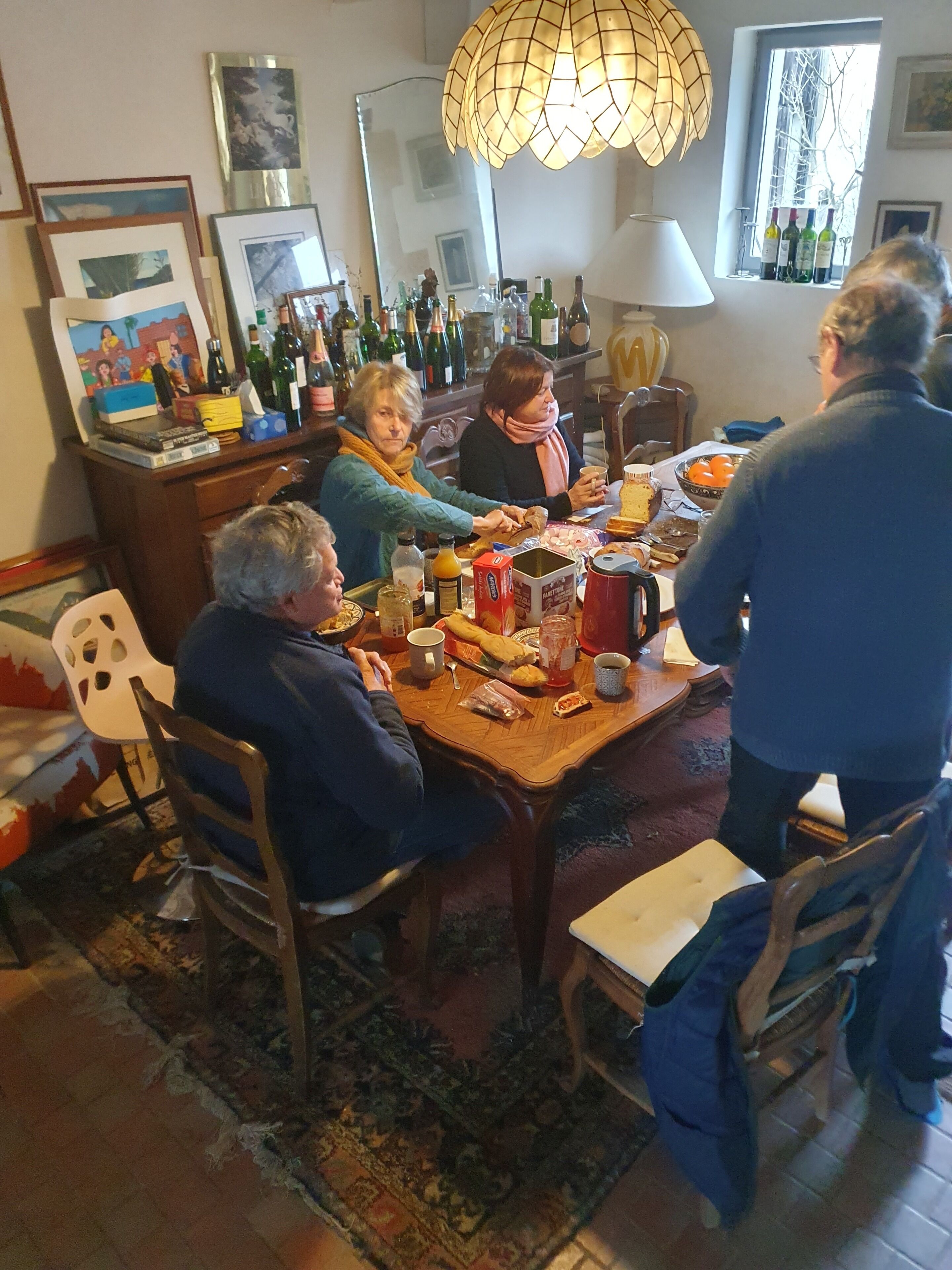 A dining area with a wooden table, chairs, and a chandelier. There are bottles and glasses on the table, and a window with bottles in the background.