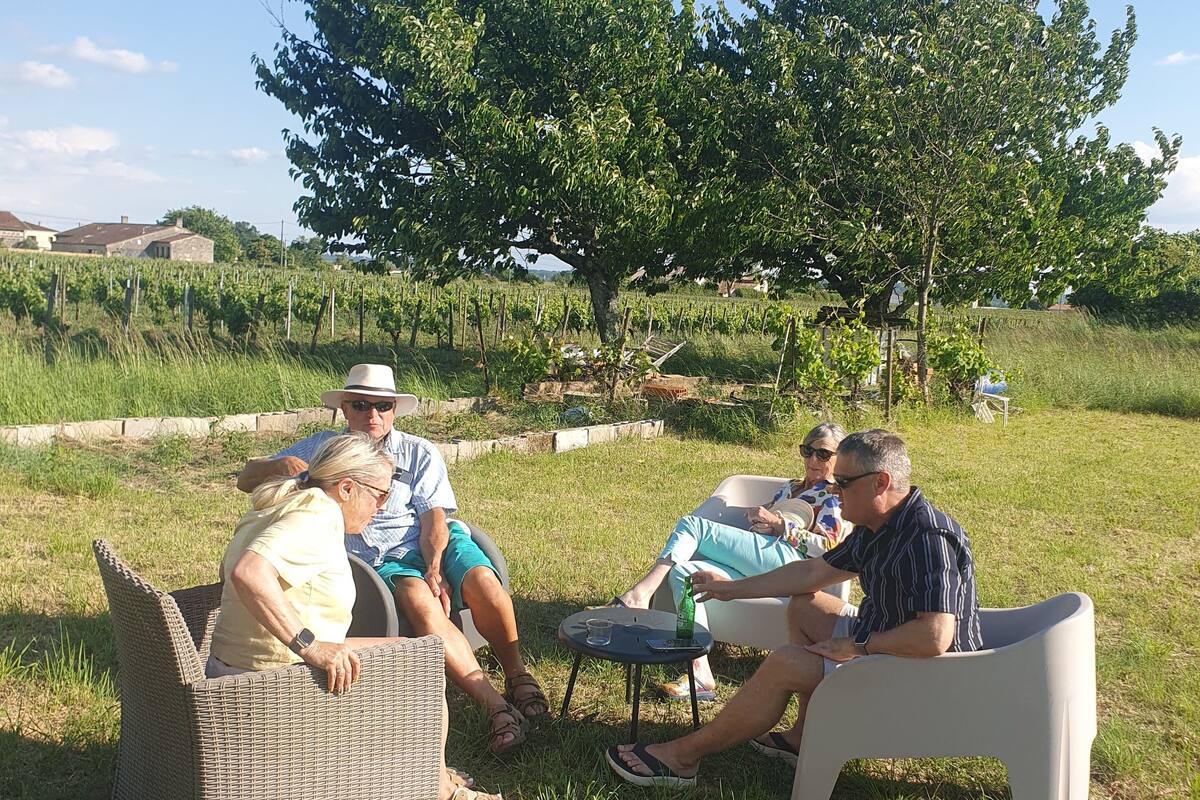 A group of people seated on outdoor furniture in a grassy area with a vineyard and trees in the background.