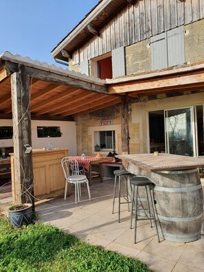 An outdoor patio area with a wooden table and chairs, a bar counter, and a large barrel. - Au Vélo dans l'Arbre (Saint-Émilion)