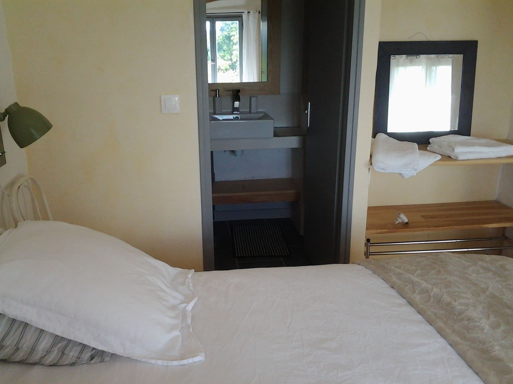 A bedroom with a bed, a bedside table, a mirror, a window, and a bathroom with a sink and towel rack.