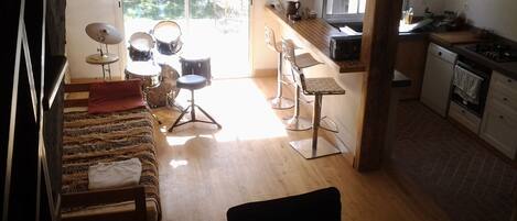 A compact living space with a sofa, a chair, a drum set, and a kitchen area.