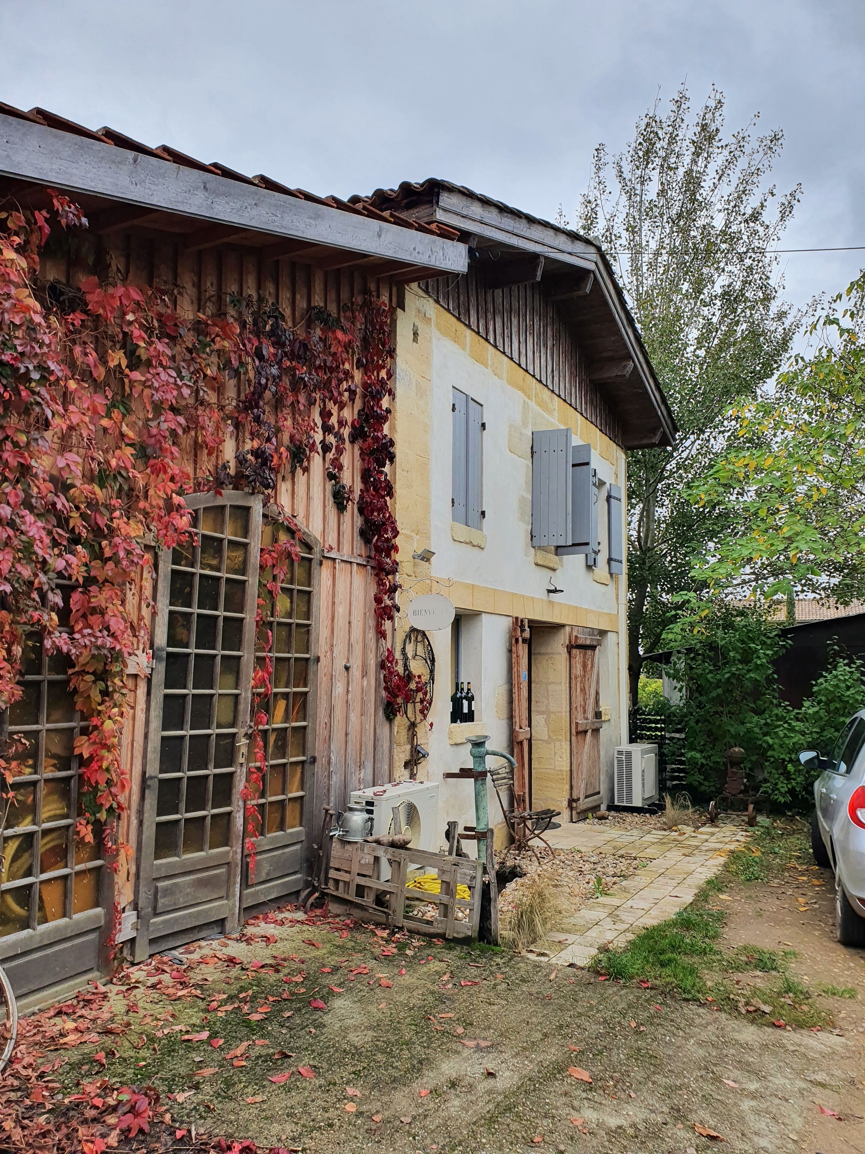 A rustic building with ivy-covered walls, a wooden door, and a small garden area with a car parked nearby.