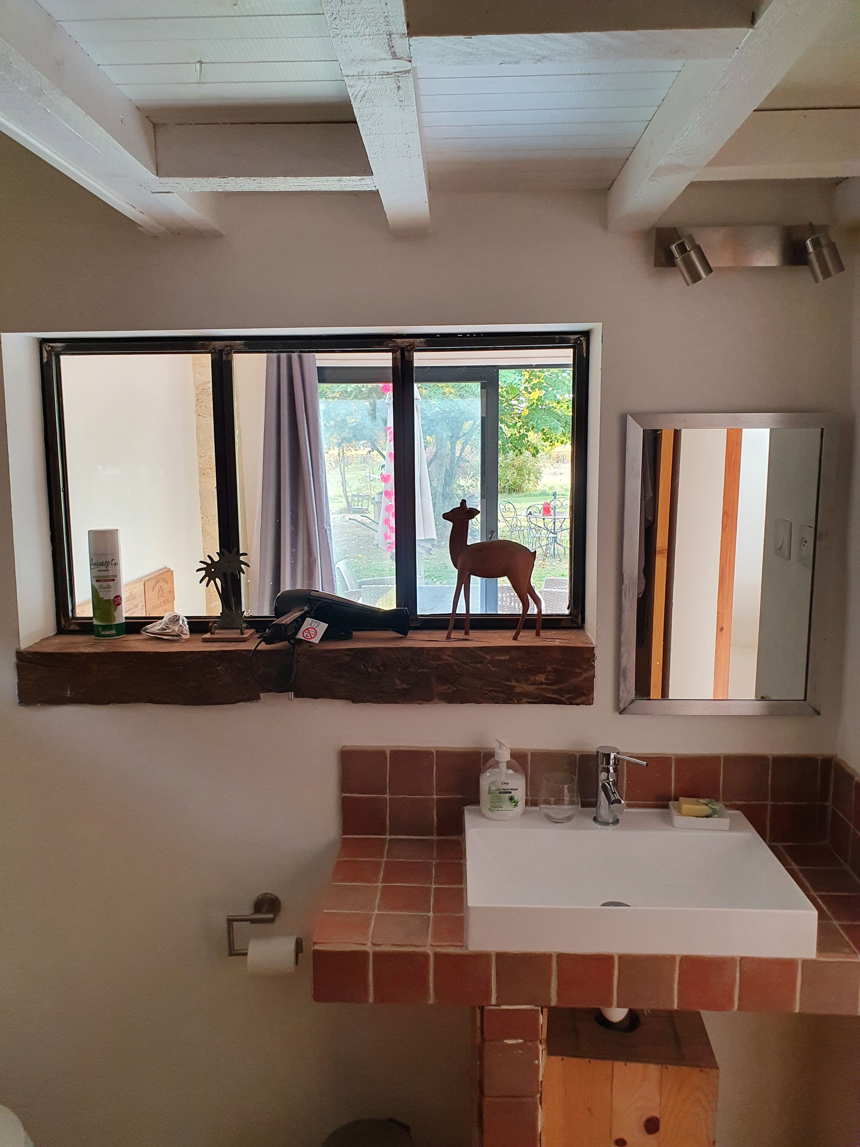A bathroom with a large window, a wooden shelf with a deer figurine, a sink with a soap dispenser, and a mirror.