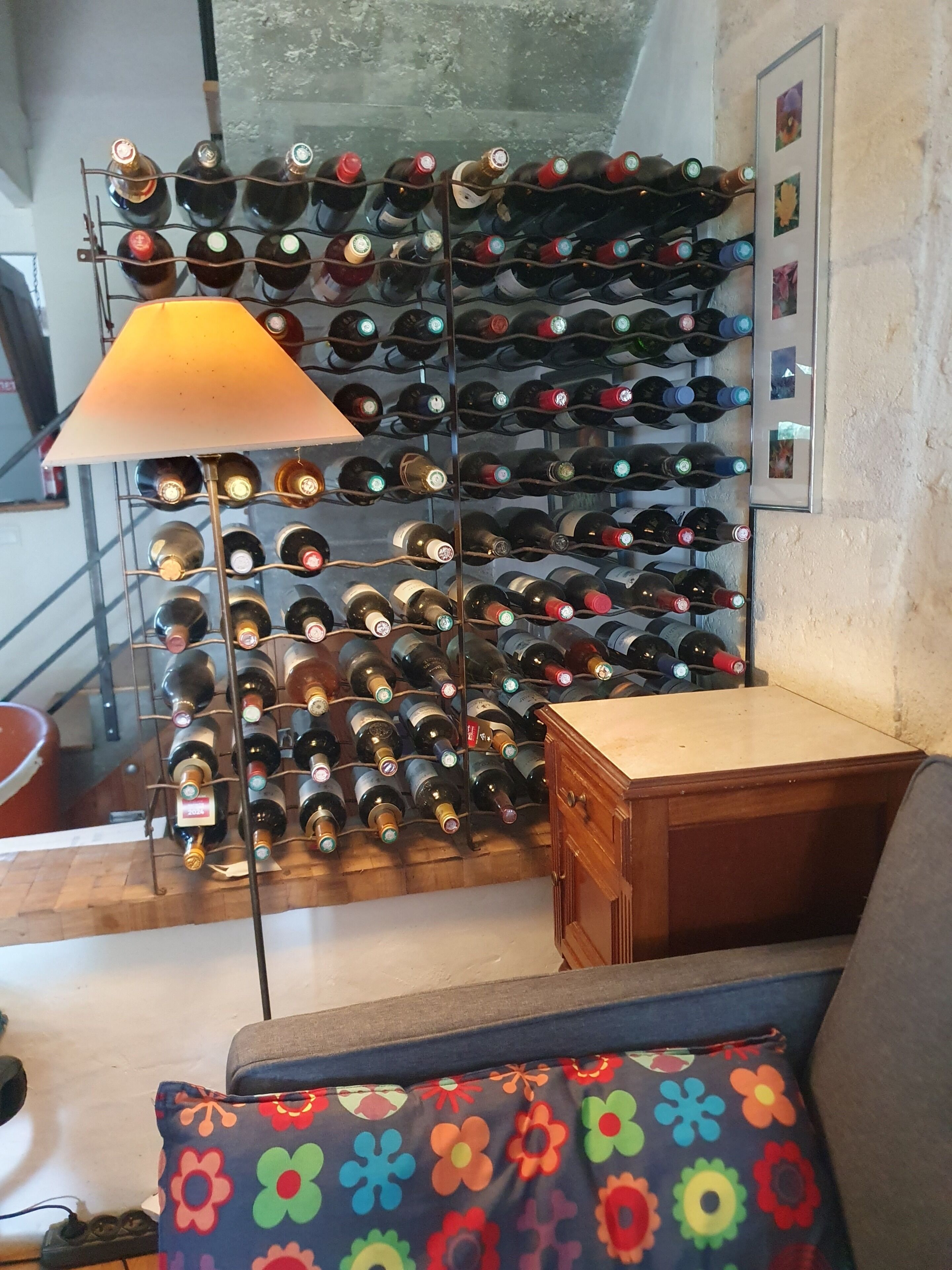 A wine rack with multiple bottles, a lamp, and a wooden cabinet in a room with a patterned cushion.