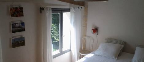 Un dormitorio con cama, ventana con cortinas, una silla y cuadros en la pared.
