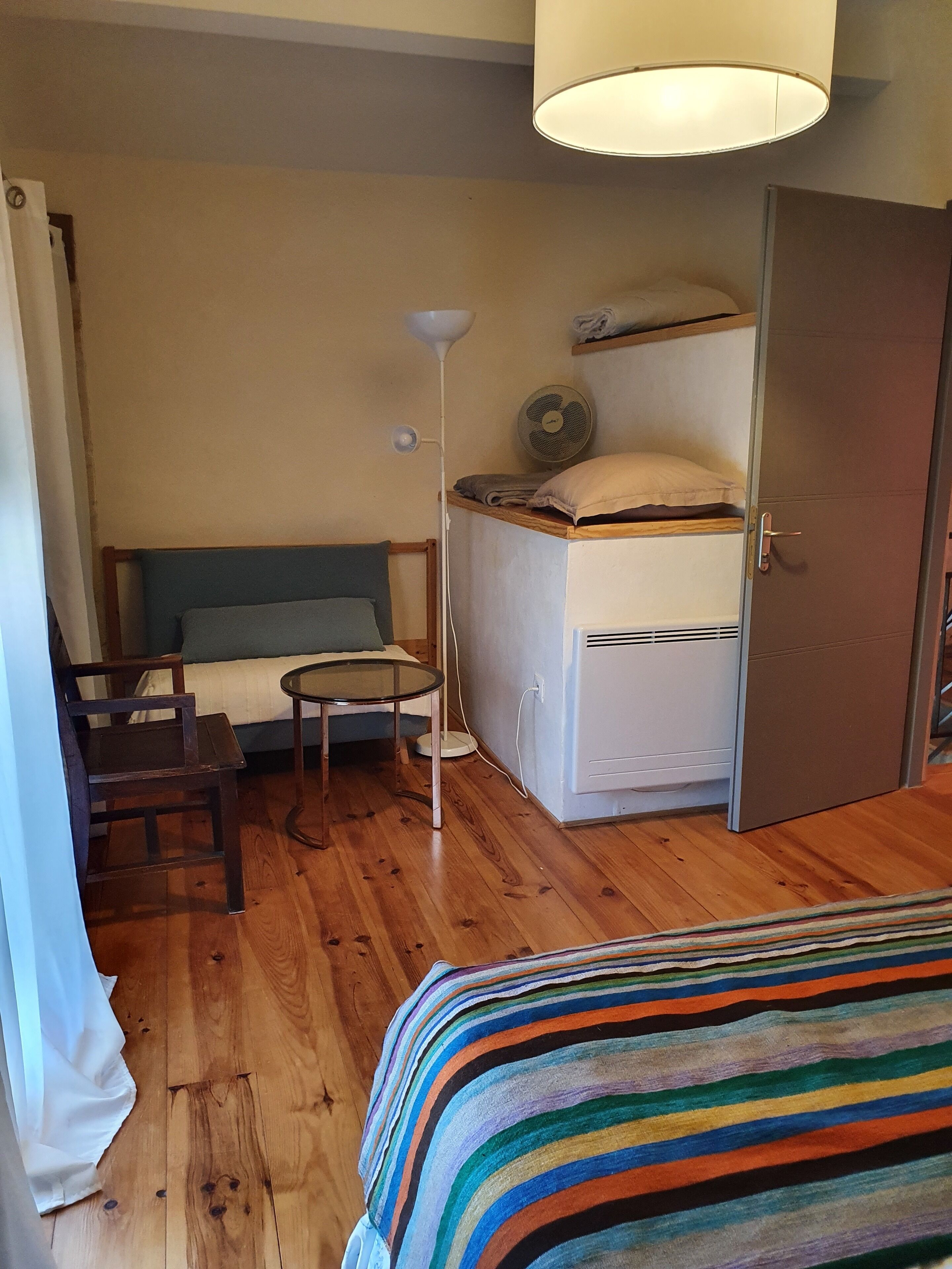 A compact hotel room with a bed, a small table, a chair, a bedside table, a lamp, a fan, and a wardrobe.