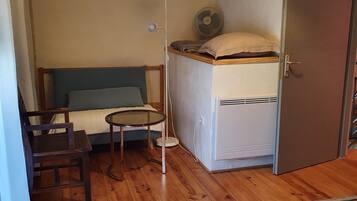 A compact hotel room with a bed, a small table, a chair, a bedside table, a lamp, a fan, and a wardrobe.