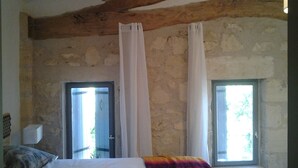 Un dormitorio con pared de piedra, viga de techo de madera, cama con manta colorida y dos ventanas con cortinas.