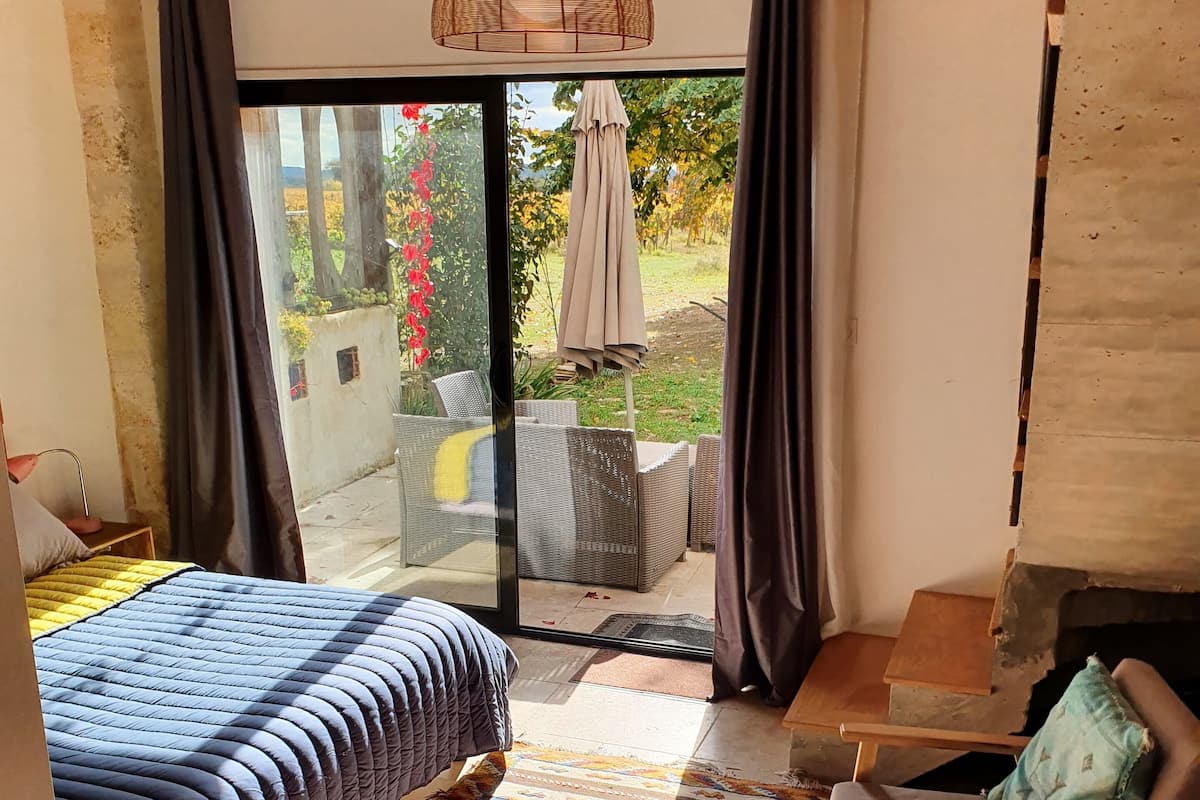 A bedroom with a large bed, a small table, a chair, and a view of the garden through a glass door.