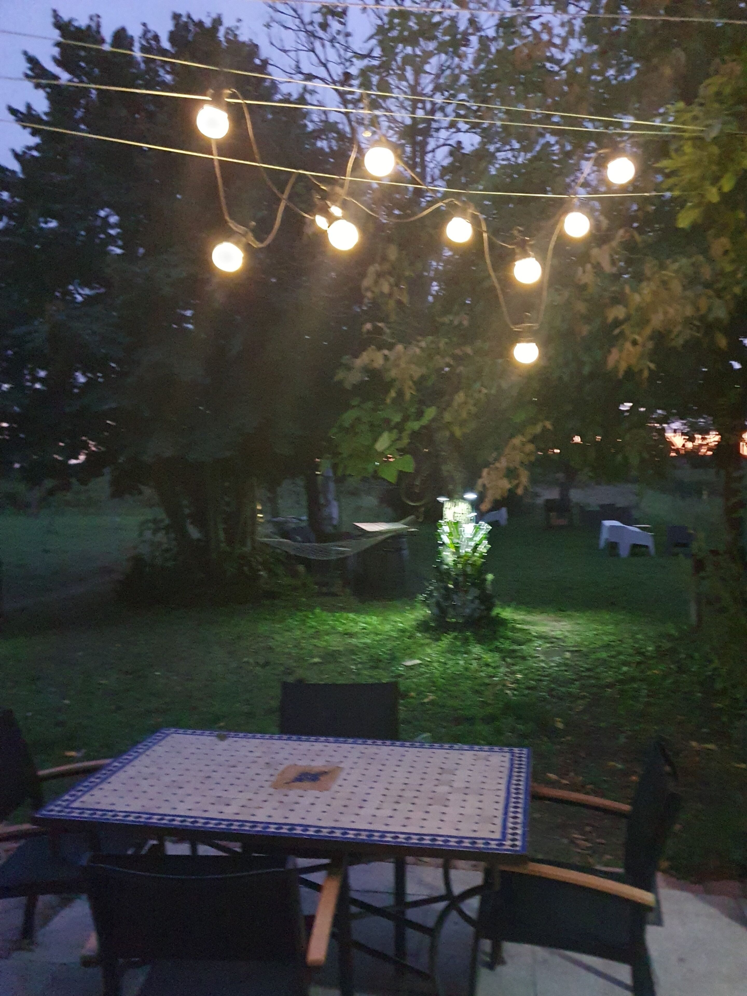Un patio al aire libre con una mesa y sillas, luces colgantes y un área con césped y árboles.