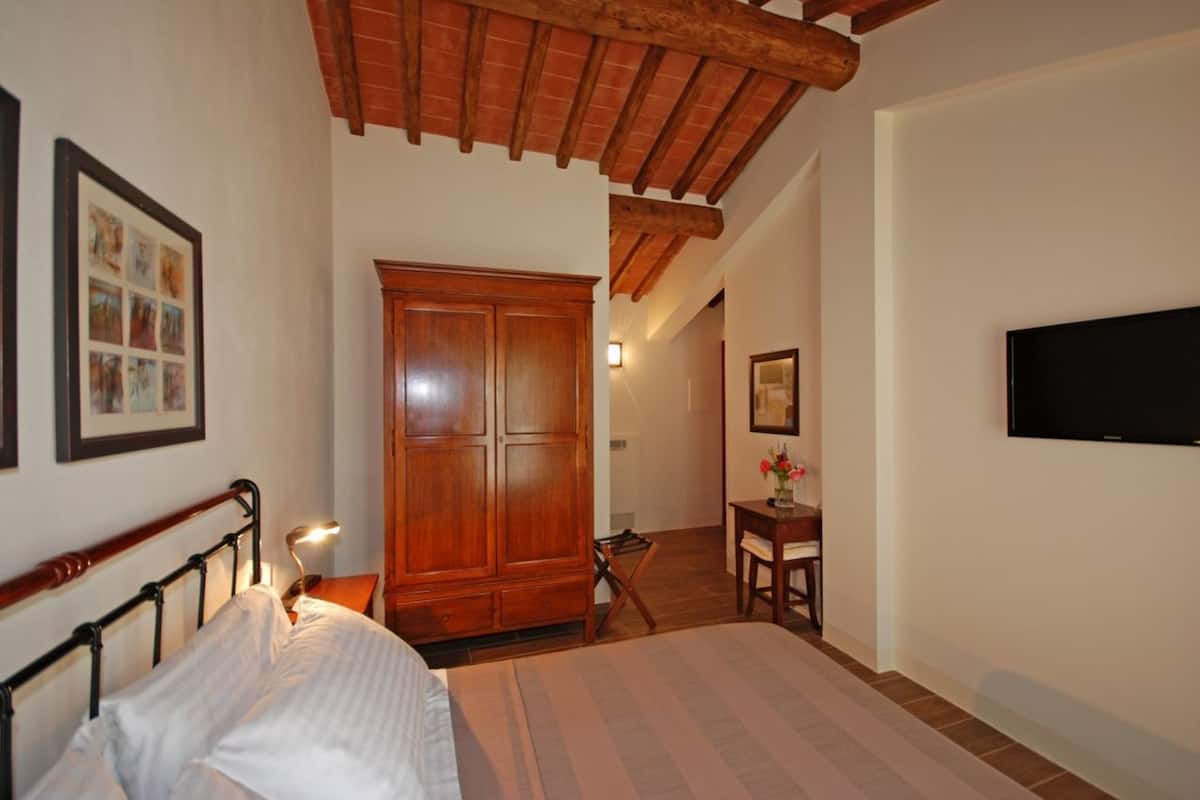 A bedroom with a wooden bed, a wardrobe, a desk with a chair, and a television on the wall.