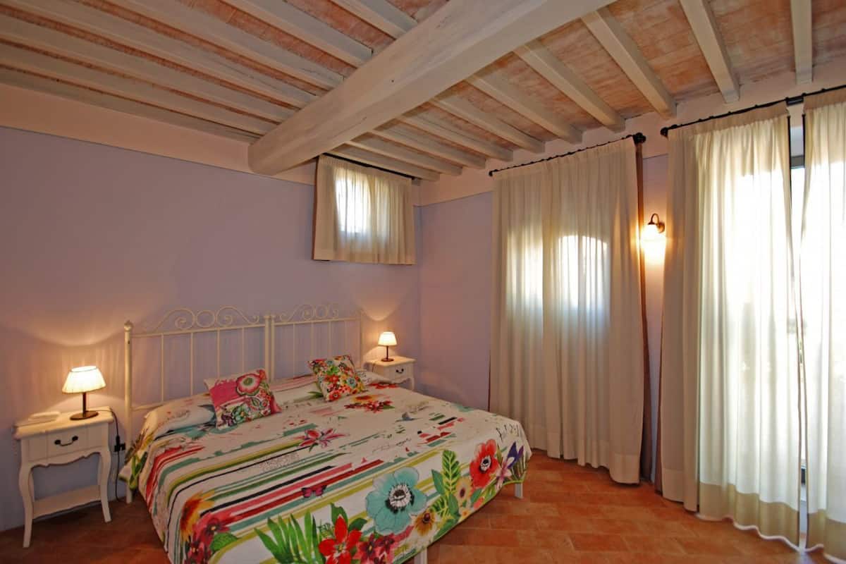 A bedroom with a bed, two bedside tables with lamps, a window with curtains, and a wooden ceiling.