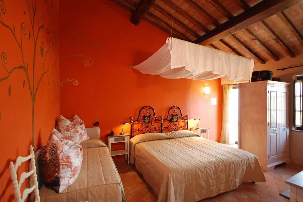 A bedroom with two beds, a canopy bed, orange walls, wooden ceiling beams, a wardrobe, and a nightstand with a lamp.