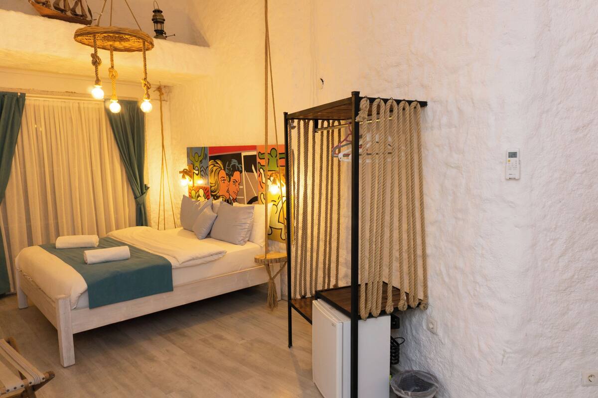 A bedroom with a bed, a small table, a hanging chair, and a shelf with a lamp.