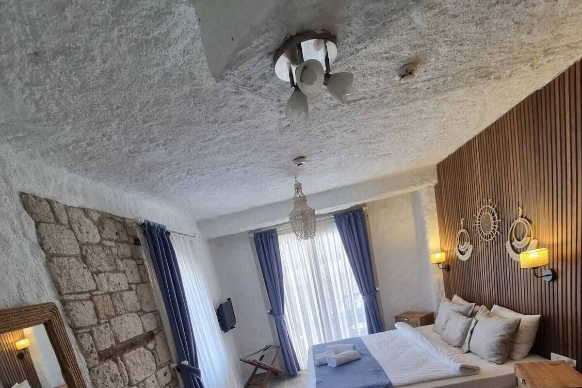 A bedroom with a stone wall, a bed with a blue bedspread, a wooden nightstand, a wooden table, a hanging light fixture, and patterned floor tiles.