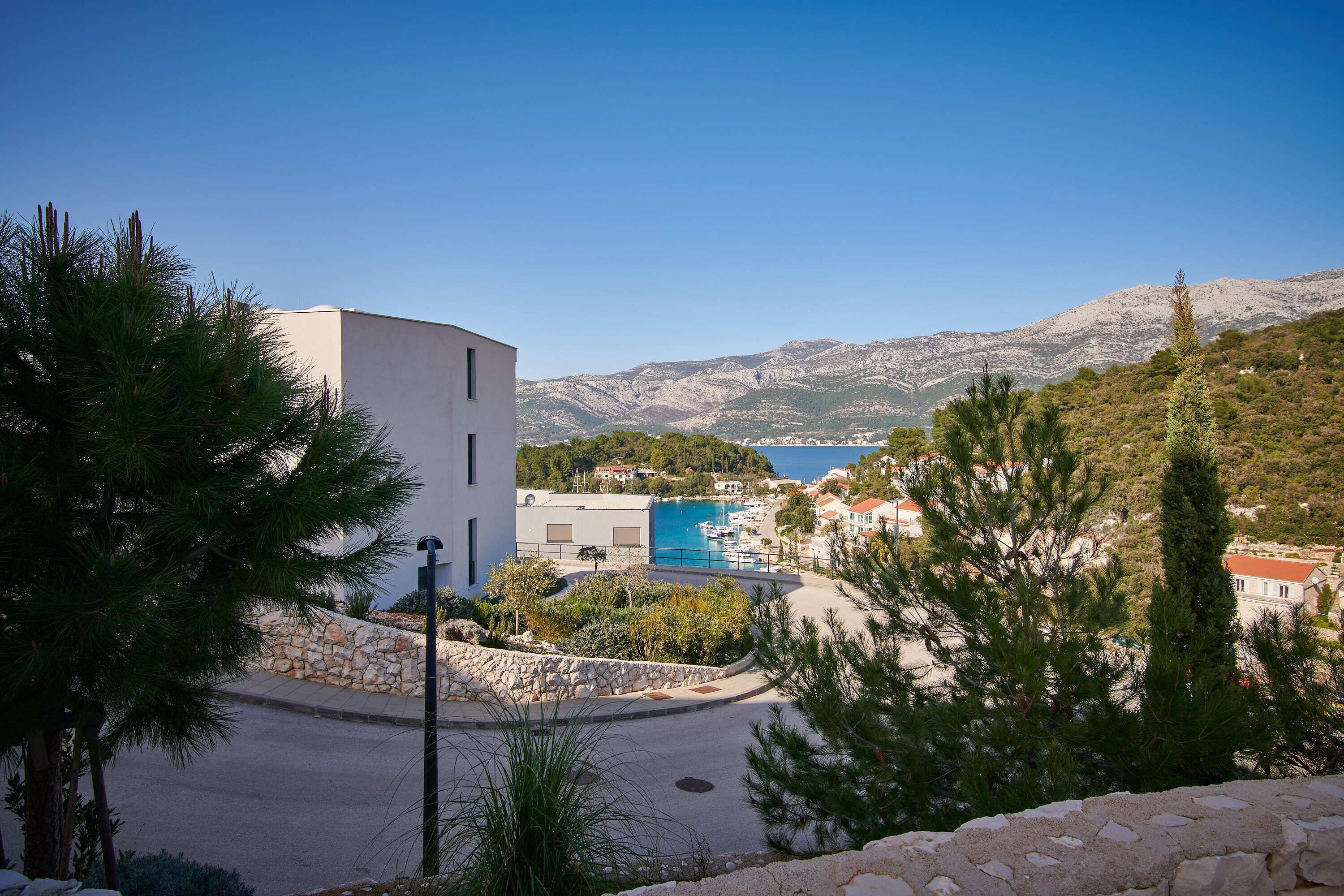 A coastal building with a stone wall, a clear blue sky, and a view of the sea and mountains in the background.