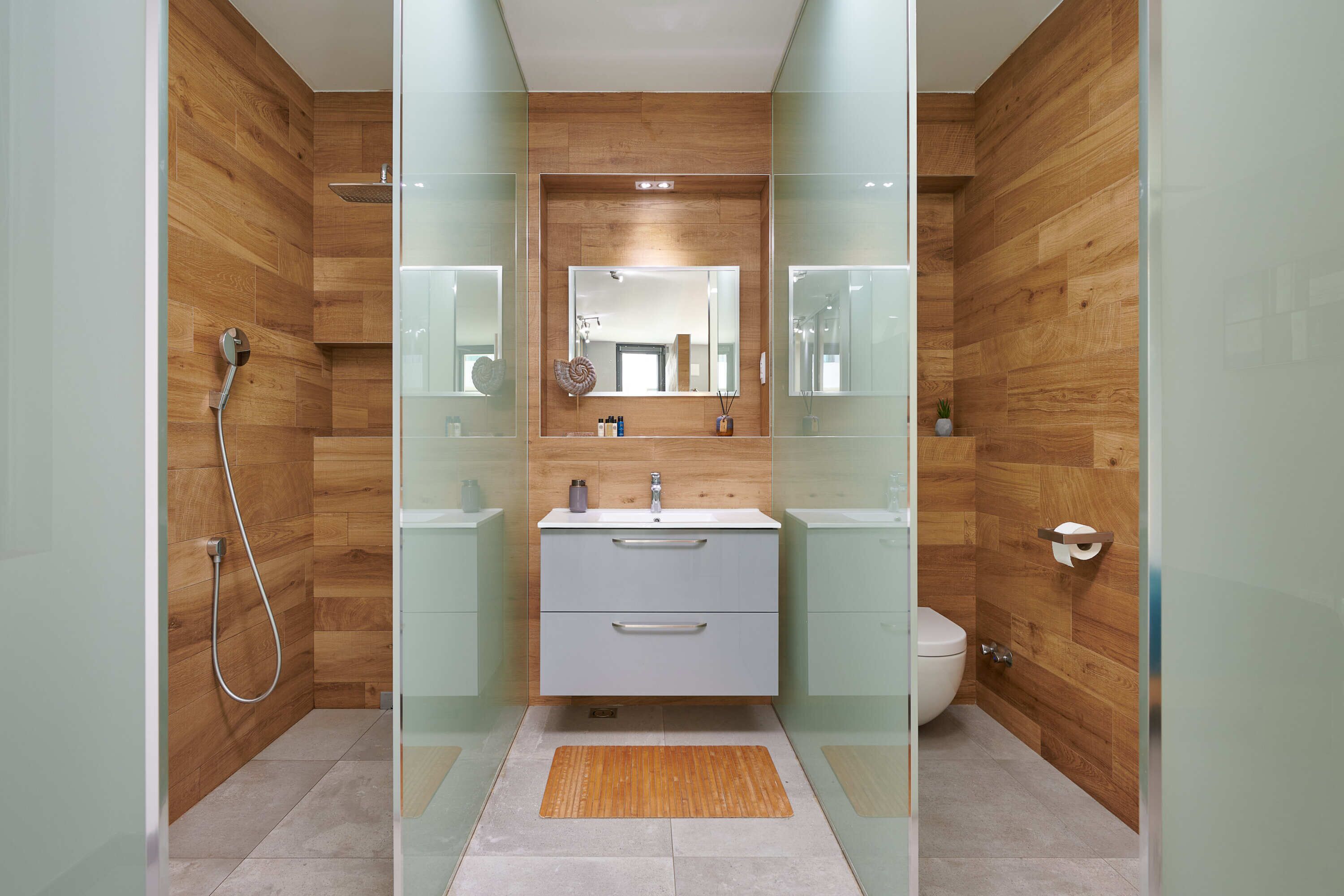 Un baño moderno con paneles de madera, cabina de ducha de vidrio, lavamanos blanco con espejo arriba y retrete con pared de paneles de madera.