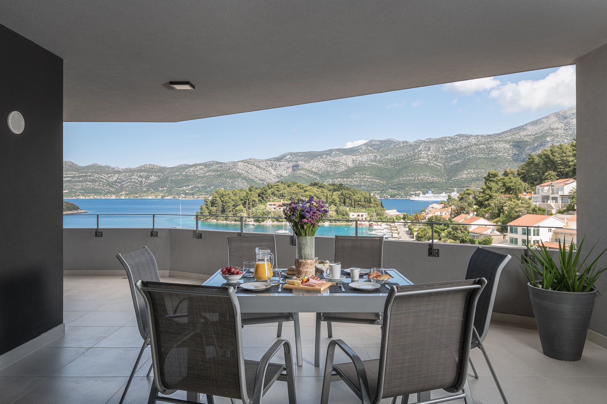 Ein Balkon mit einem Frühstückstisch, der Blick auf die Küste mit Bergen und Häusern freigibt.